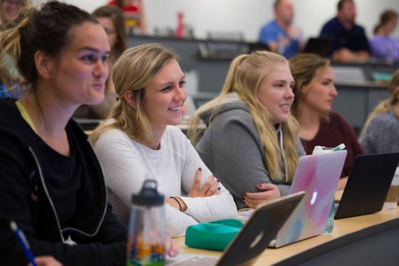 Photo of four female Chatham University students smiling in a lecture hall, with their laptops open in front of them