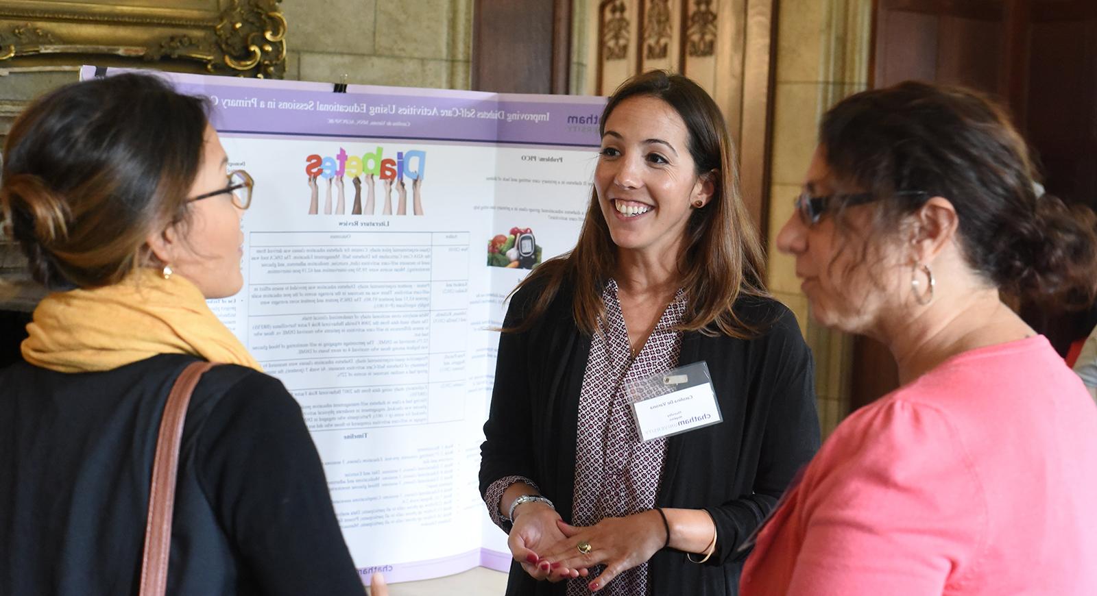 Carolina De Varona speaks to two women wearing business casual attire in front of a poster presentation. 