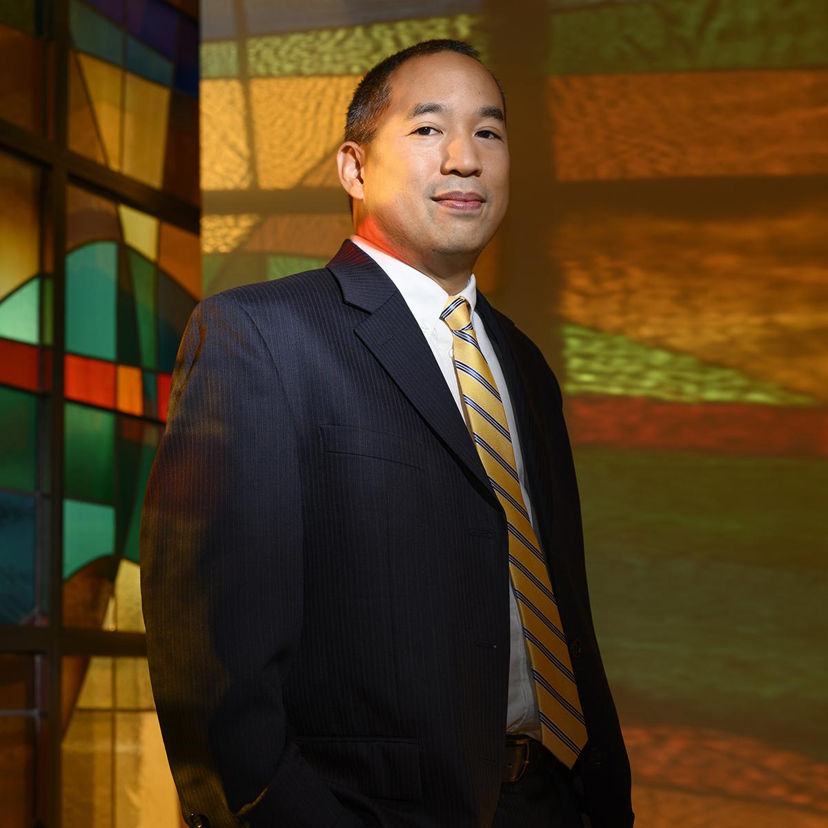 Photo of a smiling man in a suit, standing in front of colorful stained glass