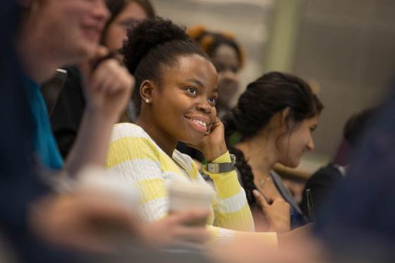 Photo of a woman in yellow sweater sitting in a lecture hall, smiling and taking notes during a class. 