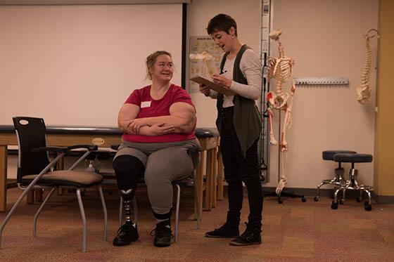 Photo of a Chatham University student looking at a clipboard and assisting a seated patient, who is wearing a leg prosthetic
