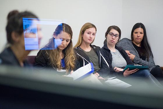 Photo of five Chatham University psychology students sitting behind glass in an exam room. There is a reflection of a screen on the glass. 