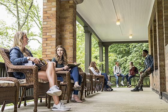 Photo of Chatham University students sitting outside on a wraparound porch, some standing and some seated in wicker chairs, talking and studying together. 