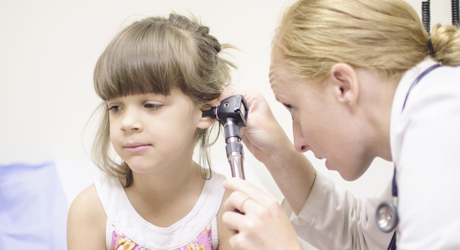 Photo of a medical professional in a white coat looks into a child's ear in an exam room.