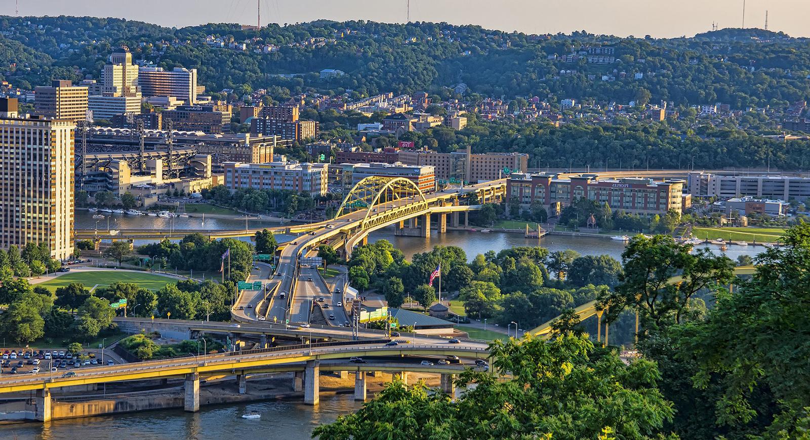 Photo of Pittsburgh's yellow bridges and rolling hills framing rivers at sunset. 