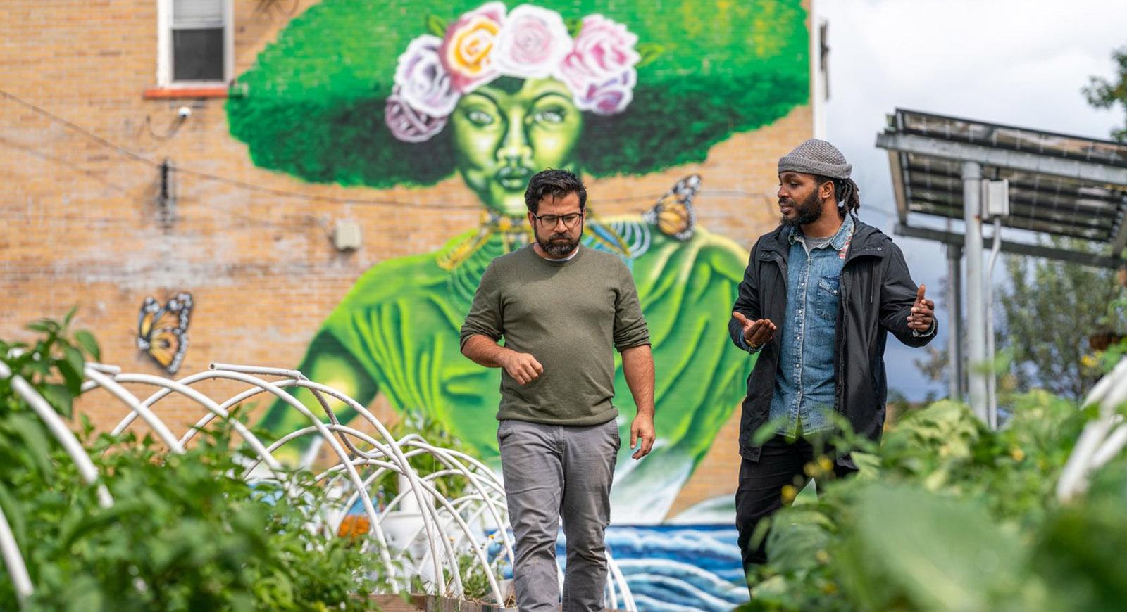 两个男人在社区花园散步聊天的照片, 有一面砖墙，背景是一幅巨大的彩色壁画
