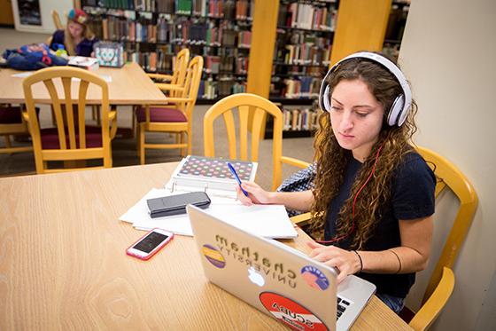 Chatham University student sits at Jeanie King Mellon Library, studying at a table while wearing headphones and working on her computer. 