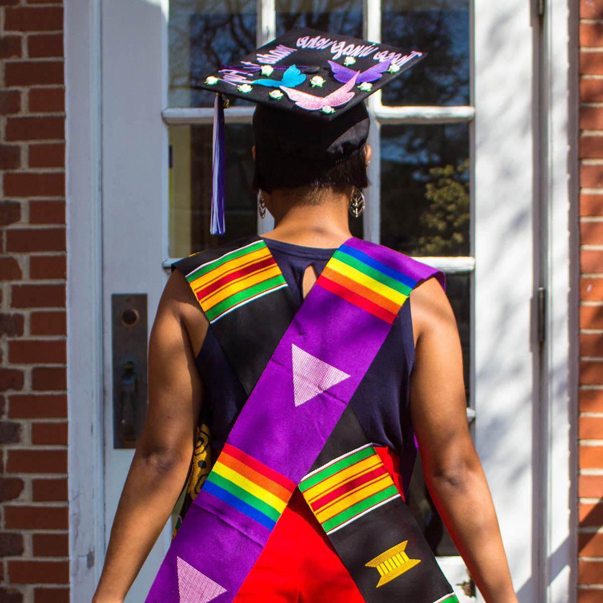 Photo of a student facing away from the camera, wearing a rainbow graduation stole and a graduation cap reading "these wings were made to fly"