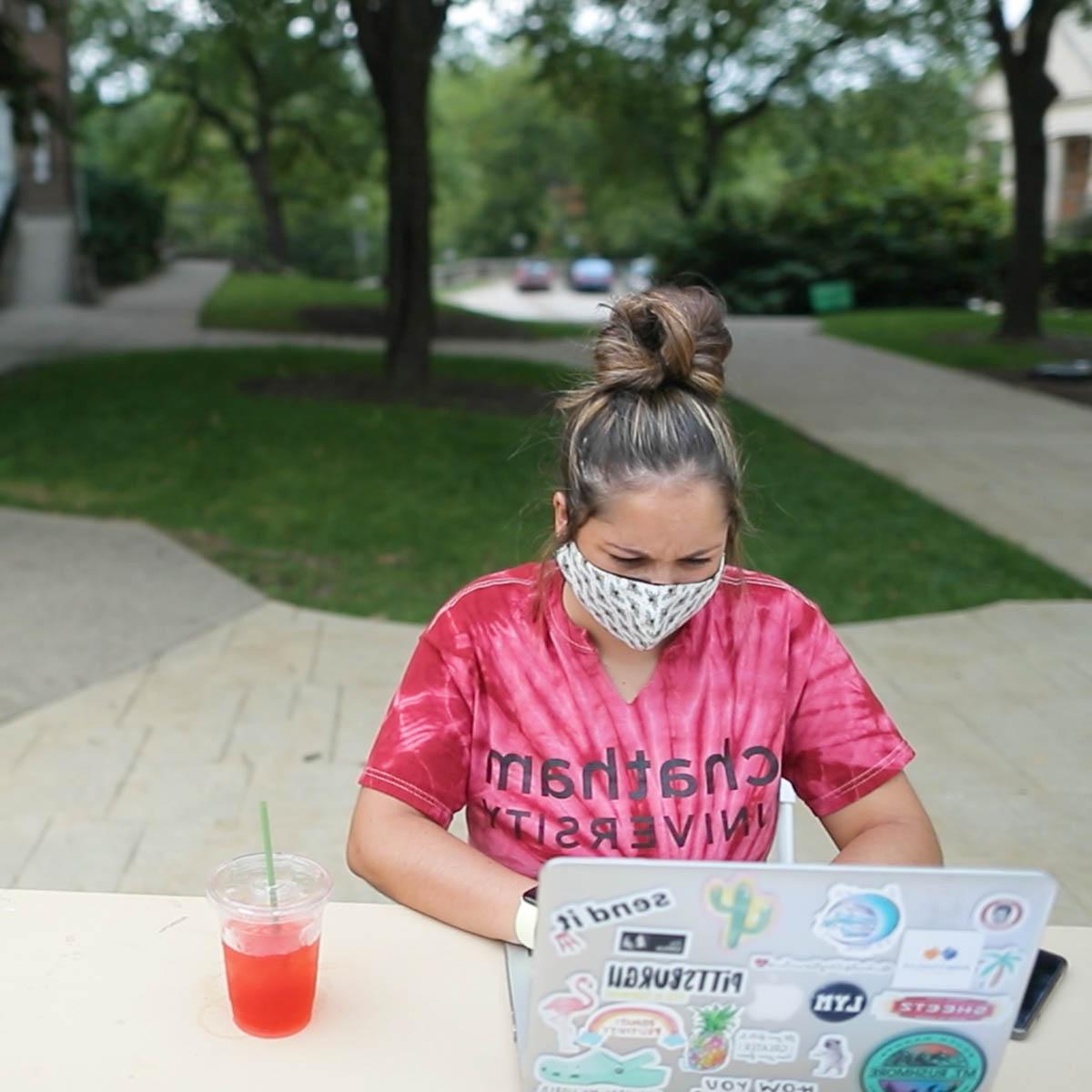 Photo of a young female student in a tie dye Chatham University shirt, wearing a mask and seated at a table in the quad on Shadyside Campus