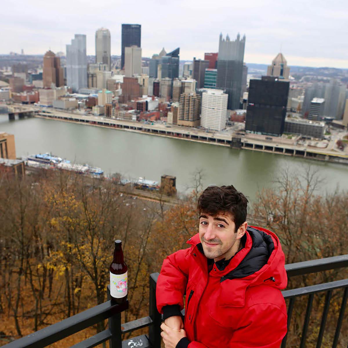 Photo of a young man in a red jacket posing with a beer bottle in front of the Pittsburgh skyline